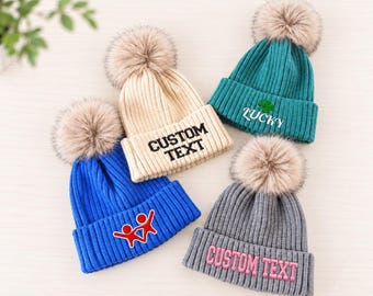 May include: Four ribbed knit beanies with faux fur pom-poms. One beige beanie reads CUSTOM TEXT in black. One teal beanie features a green clover and the word LUCKY. One blue beanie has a red graphic. One gray beanie reads CUSTOM TEXT in pink.