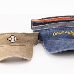 Peut inclure: Une collection de cinq visières en coton de différentes couleurs, dont beige, bleu, gris, noir et orange. La visière beige présente un motif de baseball brodé avec des étoiles, tandis que la bleue affiche le texte Custom design et un tournesol.