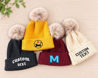 May include: Four ribbed knit beanies with faux fur pom-poms in black, yellow, maroon, and cream. The black beanie features white text reading custom text, the yellow one has a black cat and red paw print, the maroon one has a blue letter M, and the cream one has black text reading CUSTOM TEXT.