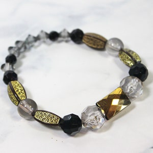 Op de afbeelding: Een armband van zwarte, gouden en heldere glazen kralen met een gouden metalen bedel. De armband is gemaakt met ronde, gefacetteerde kralen en ovale kralen met gouden accenten.