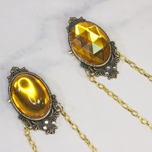 Puede incluir: Dos broches de tono dorado con piedras de vidrio amarillo de forma ovalada engastadas en marcos ornamentados. Cada broche est&aacute; unido a una cadena dorada.