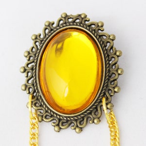 Puede incluir: Un broche ovalado dorado con una gran piedra de vidrio amarillo brillante engastada en el centro. El broche tiene un marco decorativo y adornado y dos cadenas dor&eacute;es unidas a la parte inferior.