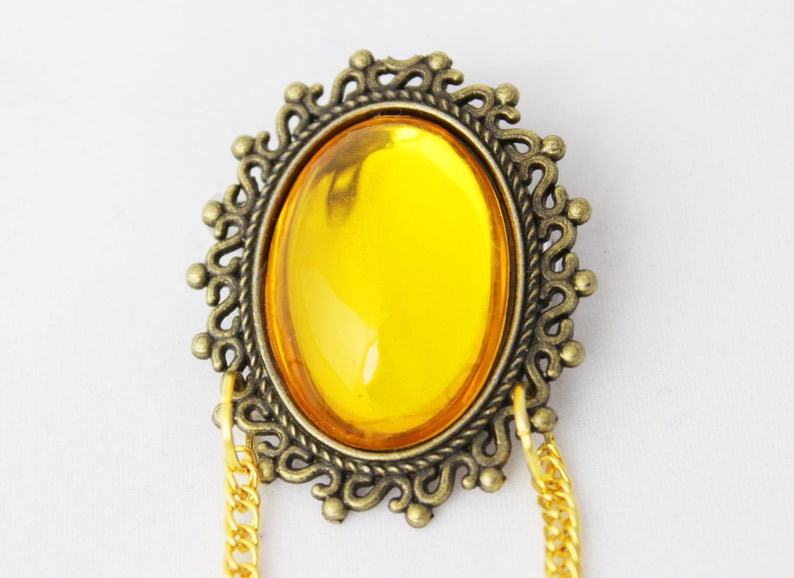Puede incluir: Un broche ovalado dorado con una gran piedra de vidrio amarillo brillante engastada en el centro. El broche tiene un marco decorativo y adornado y dos cadenas dor&eacute;es unidas a la parte inferior.