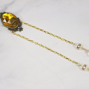 Puede incluir: Un collar de cadena dorada con un gran colgante ovalado de piedra preciosa de color &aacute;mbar. El colgante est&aacute; engastado en un marco de filigrana dorada. La cadena tiene dos gotas de cristal transparentes que cuelgan.