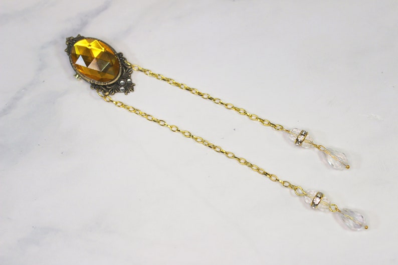 Puede incluir: Un collar de cadena dorada con un gran colgante ovalado de piedra preciosa de color &aacute;mbar. El colgante est&aacute; engastado en un marco de filigrana dorada. La cadena tiene dos gotas de cristal transparentes que cuelgan.