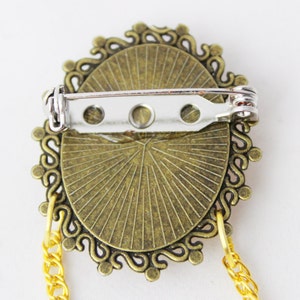 Puede incluir: Un broche de color bronce con un dise&ntilde;o decorativo y ornamentado. El broche tiene un cierre de pin y una cadena unida a la parte inferior.