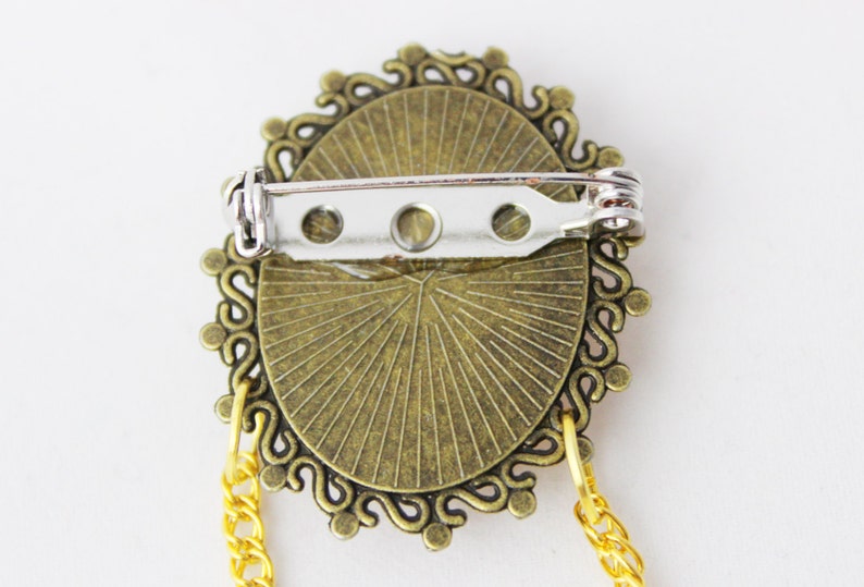 Puede incluir: Un broche de color bronce con un dise&ntilde;o decorativo y ornamentado. El broche tiene un cierre de pin y una cadena unida a la parte inferior.