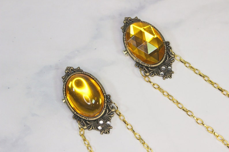 Puede incluir: Dos broches de tono dorado con piedras de vidrio amarillo de forma ovalada engastadas en marcos ornamentados. Cada broche est&aacute; unido a una cadena dorada.