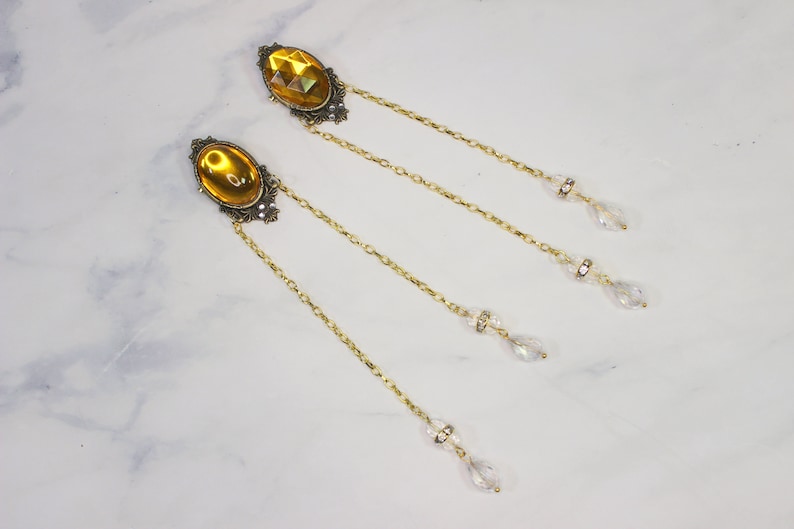 Puede incluir: Un par de pendientes de tono dorado con piedras de vidrio &aacute;mbar de forma ovalada y detalles de cristal que cuelgan de cadenas delicadas.