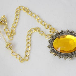 Puede incluir: Un collar de cadena dorada con un gran colgante de forma ovalada, de piedra preciosa de color &aacute;mbar, engastado en un marco adornado de color bronce.