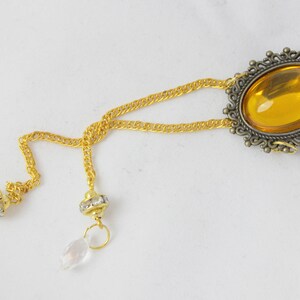 Puede incluir: Un collar de cadena dorada con un gran colgante ovalado de piedra &aacute;mbar engastado en un marco de bronce adornado. El collar tiene dos colgantes de cristal.