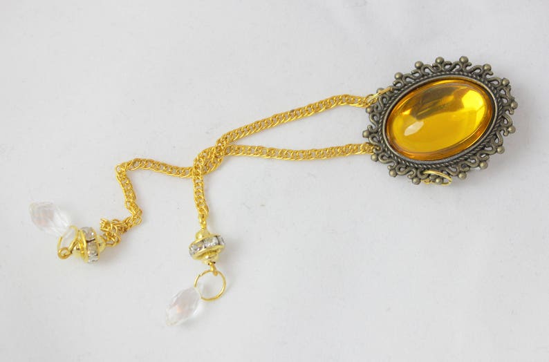 Puede incluir: Un collar de cadena dorada con un gran colgante ovalado de piedra &aacute;mbar engastado en un marco de bronce adornado. El collar tiene dos colgantes de cristal.