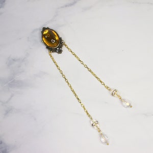 Puede incluir: Una cadena de oro con un gran colgante ovalado de color &aacute;mbar y dos abalorios de cristal en forma de l&aacute;grima.
