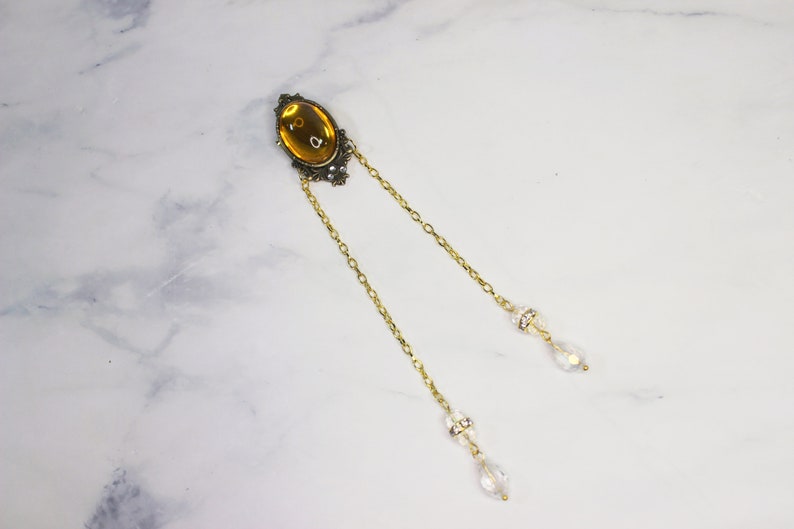 Puede incluir: Una cadena de oro con un gran colgante ovalado de color &aacute;mbar y dos abalorios de cristal en forma de l&aacute;grima.