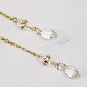 Puede incluir: Dos cadenas doradas con cuentas de cristal transparente y pedrer&iacute;a. Las cadenas cuelgan de una superficie blanca.