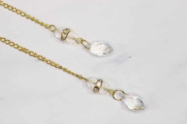 Puede incluir: Dos cadenas doradas con cuentas de cristal transparente y pedrer&iacute;a. Las cadenas cuelgan de una superficie blanca.