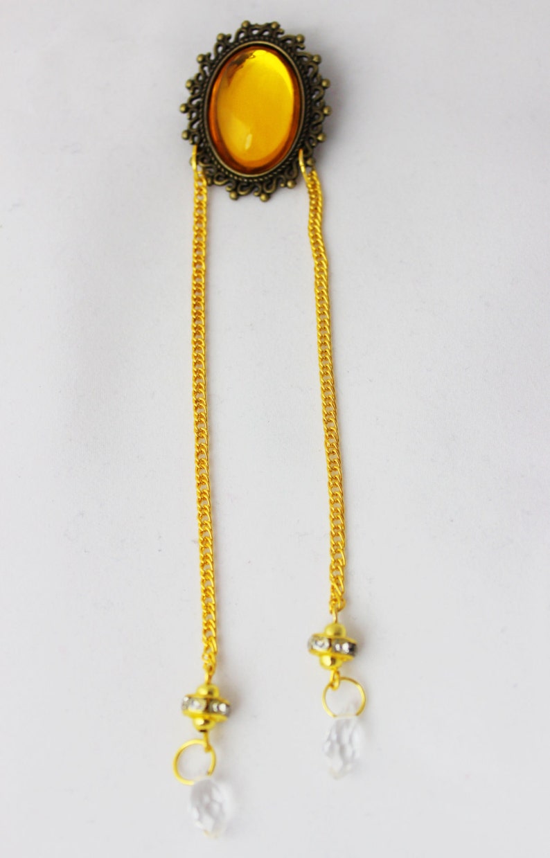 Puede incluir: Una cadena de oro con una piedra ovalada grande de color &aacute;mbar engastada en un marco de bronce decorativo. La cadena tiene dos gotas de cristal transparentes en los extremos.