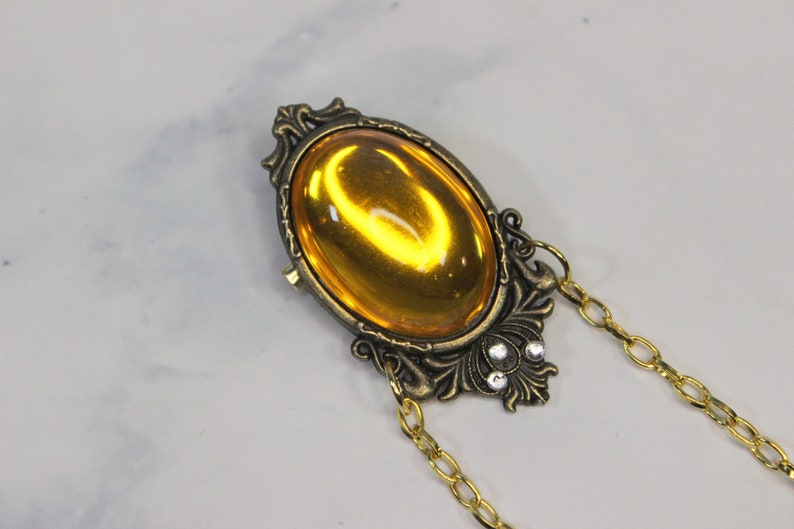 Puede incluir: Un colgante dorado con una gran piedra de vidrio de color &aacute;mbar de forma ovalada engastada en un marco adornado. El colgante est&aacute; unido a una cadena dorada.