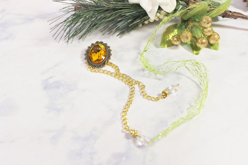 Puede incluir: Un collar de cadena dorada con un gran colgante de piedra preciosa de color &aacute;mbar. El colgante est&aacute; rodeado por un marco de metal decorativo. El collar est&aacute; colgado en una superficie blanca con una cinta verde.