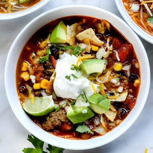 May include: A close-up shot of a white bowl filled with chili. The chili is topped with sour cream, avocado slices, shredded cheese, tortilla chips, and a lime wedge. The chili contains black beans, corn, and tomatoes.
