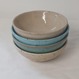 May include: A stack of four ceramic bowls with a speckled finish. The top bowl is a light beige, the second is a bright turquoise blue, the third is a muted teal green, and the bottom bowl is a speckled beige, all featuring a smooth, rounded rim.