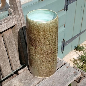 May include: A tall, cylindrical ceramic vase with a speckled brown exterior and a light blue interior. The vase is placed on a weathered wooden surface. The vase has a smooth, glossy finish. The background features a light blue door and hinges.