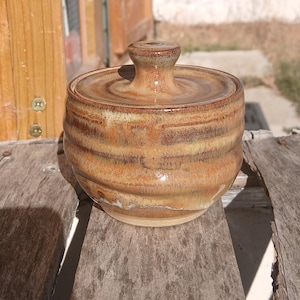 May include: A round ceramic lidded jar with a glossy finish, featuring a warm colour palette of blended brown, tan, and cream tones. The jar has a slightly ribbed texture along its sides and a small, centred knob handle on the circular lid.