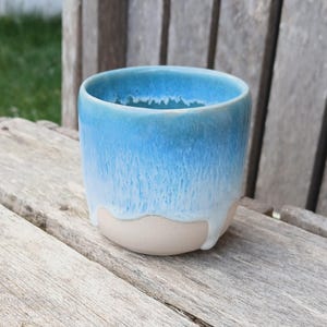 May include: A ceramic cup with a light blue glaze that fades to a creamy white at the base. The cup is cylindrical with a slightly flared rim. The glaze has a dripped effect, adding texture.