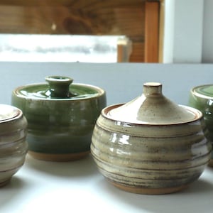 May include: Four ceramic lidded jars arranged in a group. Two jars feature a light beige glaze with horizontal brown banding, while the other two jars are finished in a solid, glossy olive green glaze. Each jar has a matching lid with a knob.