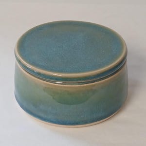 May include: A round ceramic jar with a lid. The jar is glazed in shades of blue and green, with a lighter beige rim. The jar has a smooth, glossy finish and a simple, elegant design.