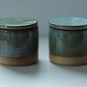 May include: Two cylindrical ceramic jars with matching lids, featuring a two-tone glaze. The upper sections are a mottled teal and blue, while the lower bases are a solid, unglazed tan clay. Each jar has a flat lid with a smooth, glossy finish.