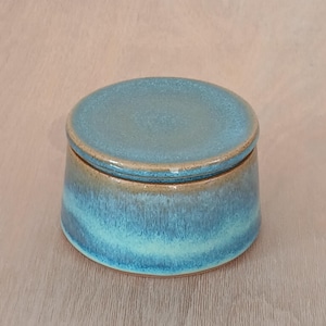 May include: A round ceramic lidded jar featuring a smooth, glossy glaze in shades of teal, turquoise, and deep blue. The container has a slightly tapered cylindrical shape with a matching circular lid that sits flush on the top rim of the vessel.