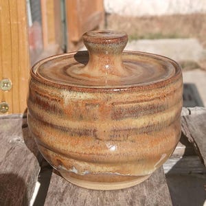 May include: A round ceramic lidded jar with a glossy finish, featuring horizontal ridges along the body. The glaze displays a blend of earthy brown, tan, and cream tones with subtle variations in colour intensity across the surface.