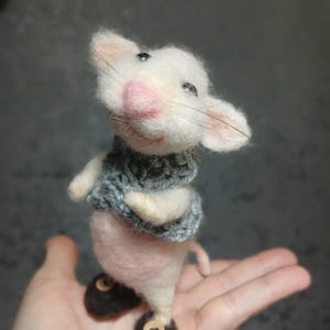 以下が含まれることがあります： 白い体、ピンクの頬、灰色のニットスカーフが付いた、手作りのフェルトマウスの置物。 マウスは、ボタンのディテールが付いた茶色のフェルトシューズを履いています。 置物は手で持たれています。