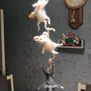 Op de afbeelding: Drie vilten muizen hangen aan een touwtje. Twee zijn wit, een met een klein oranje voorwerp, en de andere is grijs. De muizen hangen in de lucht, wat een grillige scène creëert. Een klok en andere beeldjes staan op de achtergrond.