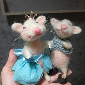 May include: Two handmade felt mice. One mouse wears a blue skirt and a gold crown. The other mouse wears a gray sweater. Both mice have pink noses and are posed as if dancing. The mice are held in a person's hand.