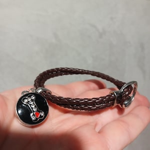 Könnte beinhalten: Ein braunes geflochtenes Lederhalsband für Hunde mit einem silbernen Verschluss. Das Halsband hat einen schwarzen runden Anhänger mit einem Knochendesign aus Strasssteinen und einem roten Herz.