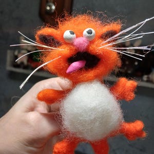 May include: A handmade orange cat figurine with a white belly, pink nose, and tongue. The cat has white whiskers and black accents on its ears. The cat's eyes are white with black pupils. The figurine is made of felt.