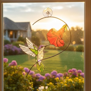 May include: A stained glass hummingbird and flower window cling. The hummingbird has a purple head, green wings, and a yellow tail. The red flower is in the shape of a trumpet. The cling is attached to a window with a suction cup.