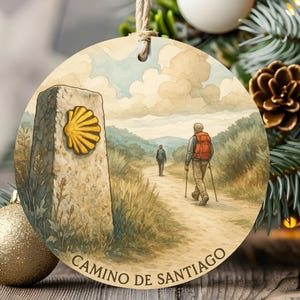 May include: A round, flat, hanging ornament featuring a watercolour-style illustration of a stone marker with a yellow scallop shell symbol, a path, two hikers with backpacks and walking poles, and the text CAMINO DE SANTIAGO at the bottom.