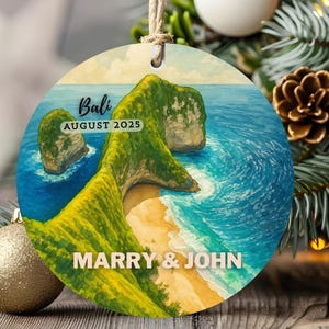 Puede incluir: Un adorno redondo de cerámica con una ilustración en acuarela de un acantilado verde sobre una playa de arena y un océano azul. El texto dice Bali, AUGUST 2025 y MARRY & JOHN. Un lazo de yute está unido a la parte superior para colgarlo.