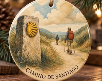 May include: A round ceramic ornament featuring a watercolor illustration of a stone marker with a yellow scallop shell, a path through a grassy landscape, two hikers with backpacks, and the text CAMINO DE SANTIAGO printed at the bottom.