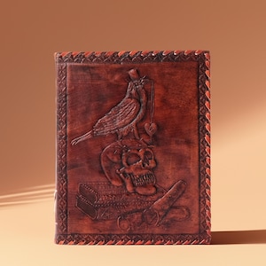 Könnte beinhalten: Ein braunes Lederjournal mit einem detaillierten Schnitzmuster. Das Cover zeigt ein Totenkopf-, Vogel- und Herzmotiv. Das Journal hat einen dekorativen Rand und ist vor einem neutralen Hintergrund platziert.