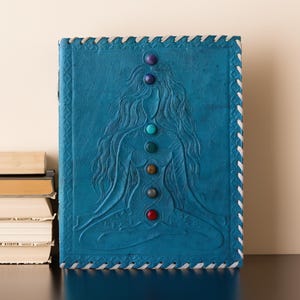 May include: A blue leather journal with a woman in a yoga pose embossed on the cover. The woman has seven gemstones embedded in her body, representing the seven chakras.