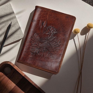 Peut inclure: Un journal en cuir marron avec un motif floral en relief. Le journal rectangulaire est fermé, présentant une image détaillée d'une fleur et de feuilles sur sa couverture. Un crayon et un bloc-notes sont visibles en arrière-plan, ainsi qu'un plateau en bois et des fleurs séchées.