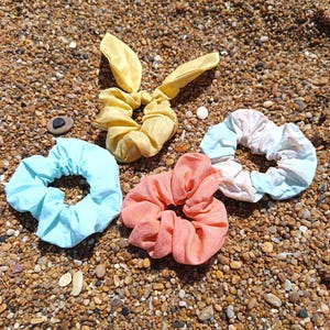 Puede incluir: Una colección de cuatro coleteros de tela. Uno es amarillo sólido con dos orejas de tela largas, uno es azul claro sólido, uno es rosa coral sólido y uno presenta un patrón tie-dye con secciones en azul claro y blanco.