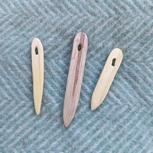 Op de afbeelding: Drie houten naalden met spitse punten en langwerpige ogen. Twee naalden zijn lichtbeige van kleur, terwijl de middelste naald een donkerder, gestreept houtnerfpatroon heeft. Elke naald heeft een gladde, taps toelopende vorm voor handwerk.