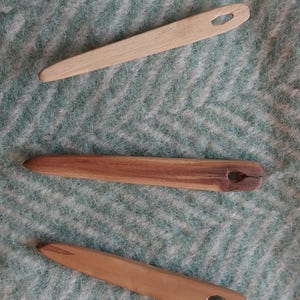 May include: Three wooden yarn needles in shades of brown and tan are displayed on a textured, light teal surface. Each needle has a pointed end and a small oval hole. These needles are likely used for weaving or other fibre arts.