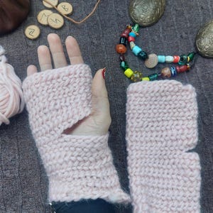 Puede incluir: Un par de guantes sin dedos de punto rosa claro. Los guantes están hechos de un hilo grueso con un punto texturizado. El diseño incluye una abertura para el pulgar y un puño. Los guantes se exhiben sobre una superficie gris.