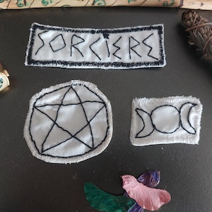Peut inclure: Divers objets mystiques, dont des écussons brodés avec des coutures noires sur tissu blanc. L'un des écussons porte l'inscription "SORCIERS". D'autres présentent un pentagramme et un motif de triple lune. Une bague dorée avec un motif floral est également présente.